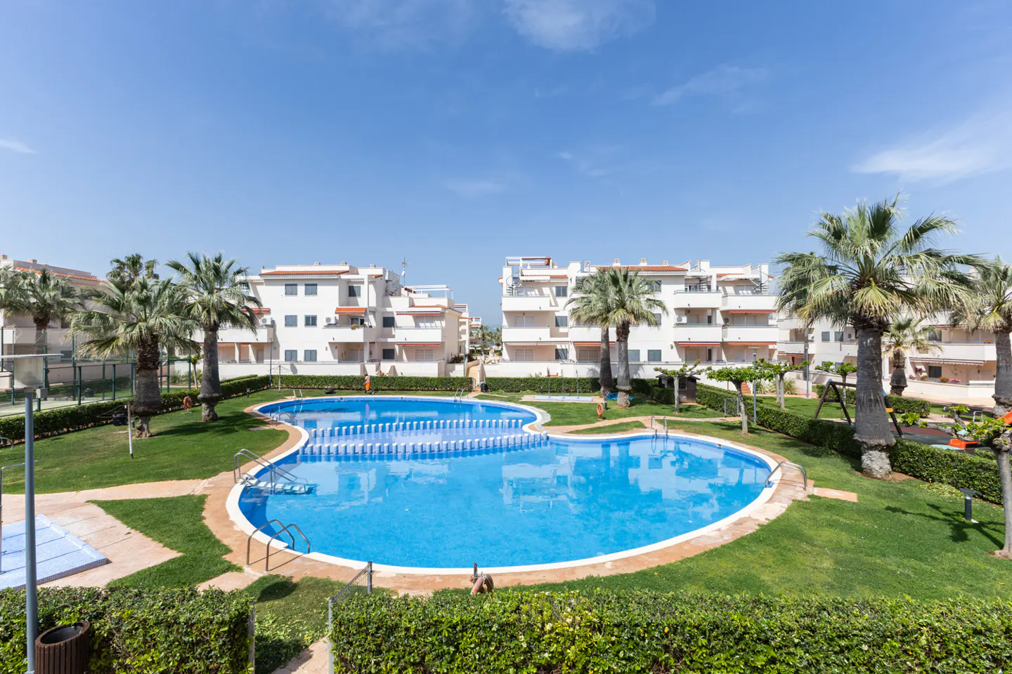 A bright blue swimming pool sits in a green lawn, surrounded by white apartment buildings and palm trees under a blue sky.