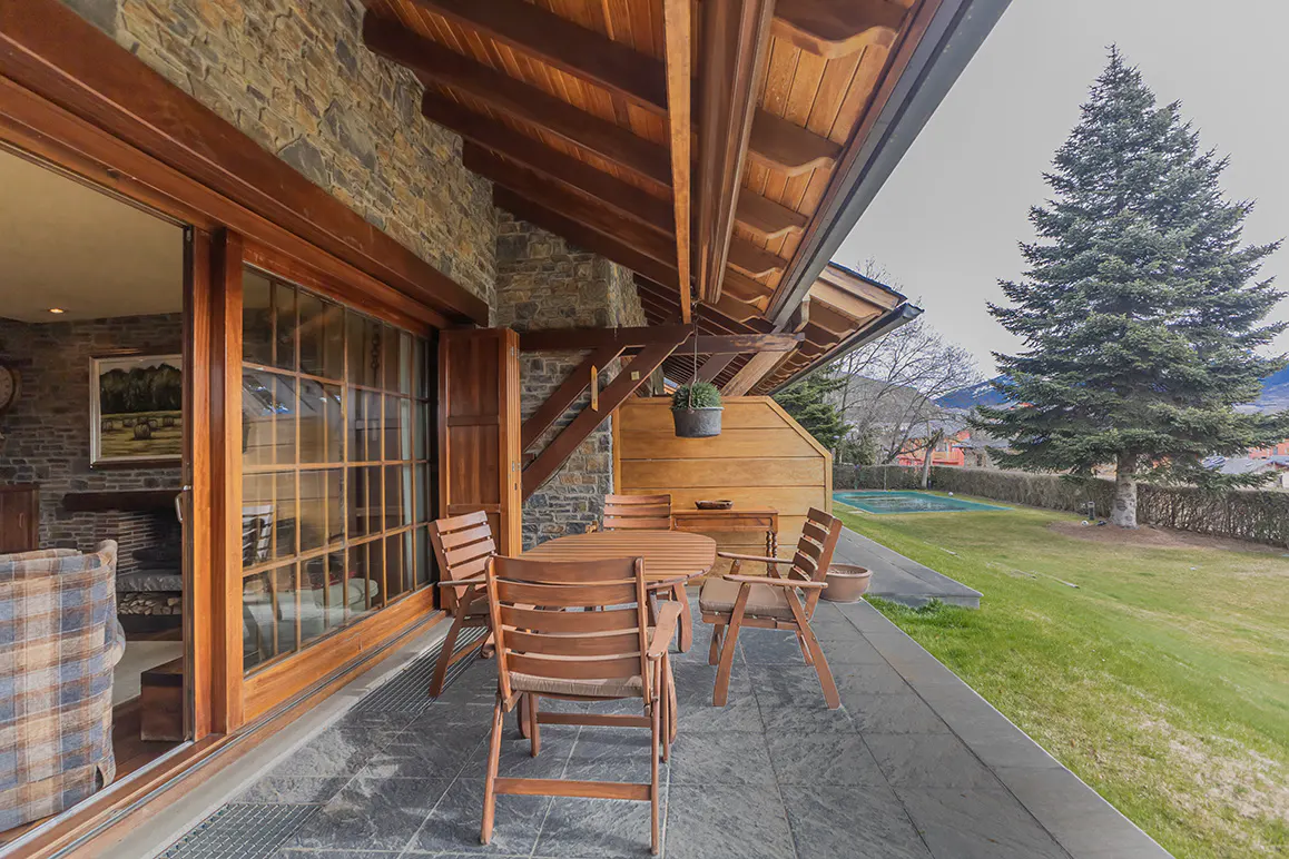 Outdoor patio with wood table and chairs on stone tiles, next to a stone wall and sliding glass doors. A green lawn and trees are in the background.