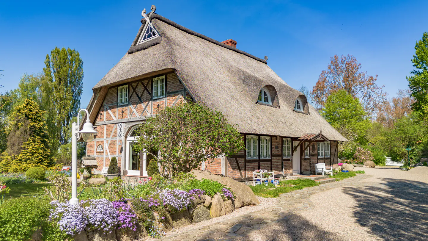 Charming half-timbered cottage with a thatched roof, surrounded by a lush garden and a gravel path under a clear blue sky.