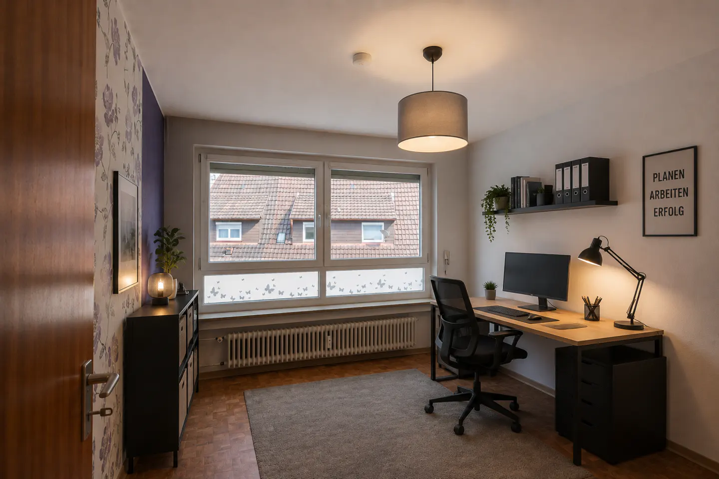 A home office with a desk, computer, and black chair. A window overlooks rooftops. A gray rug covers the floor. A black cabinet is on the left.