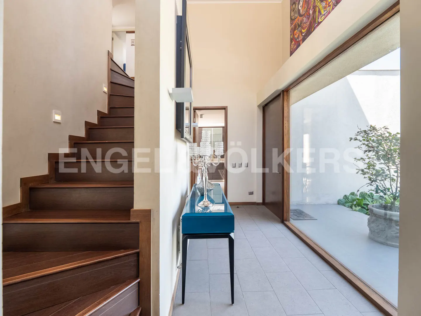 Interior view of a home's foyer with a wooden staircase, blue console table, and a large window overlooking a courtyard.