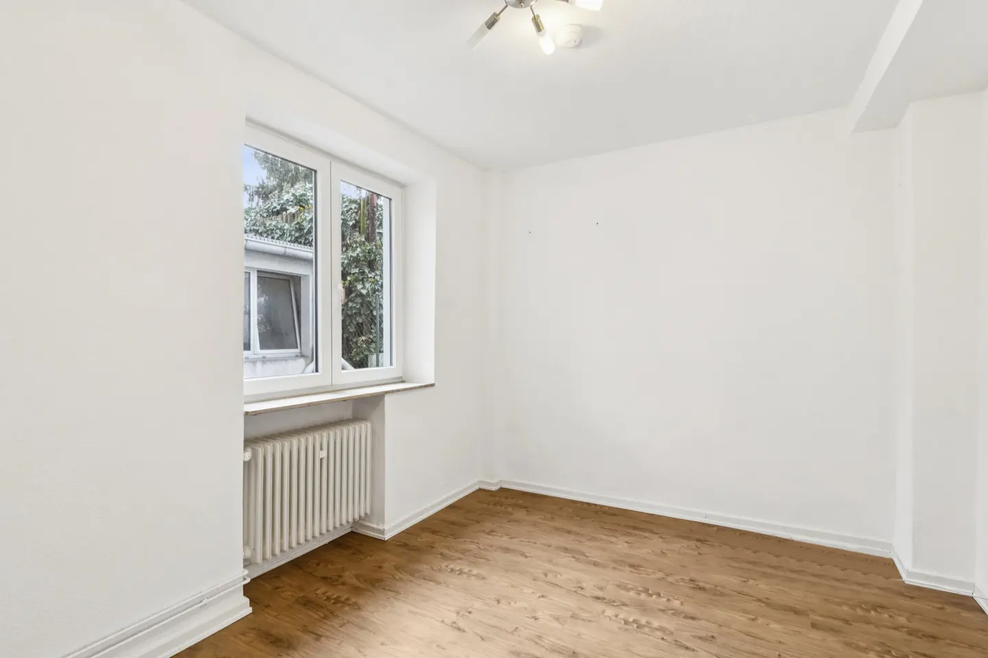 Bright, empty room with wood floors, white walls, and a window showing trees outside. A radiator sits beneath the window.