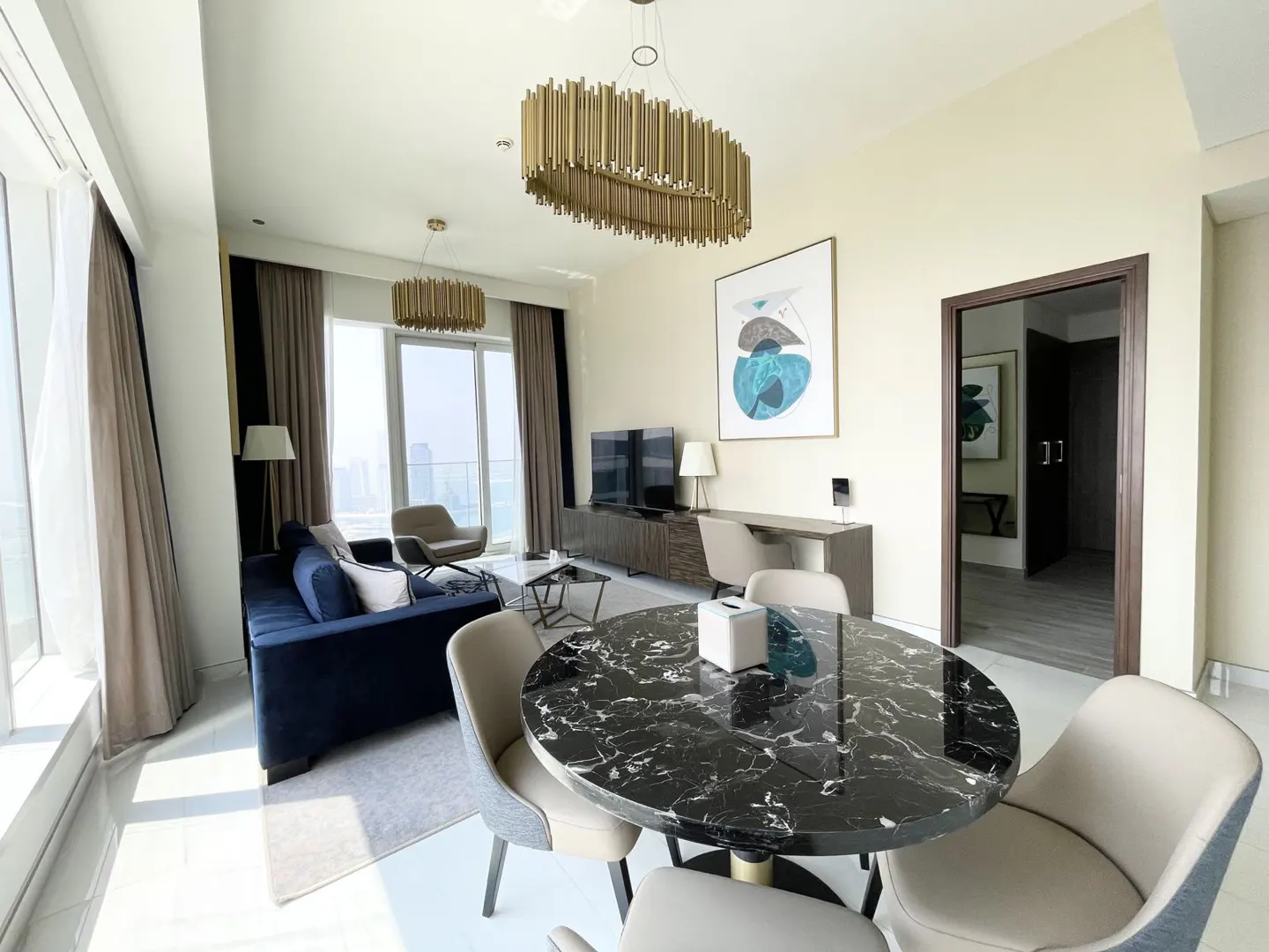 Bright, modern apartment interior with a blue sofa, marble table, and city view from large windows. Gold chandeliers add a touch of elegance.