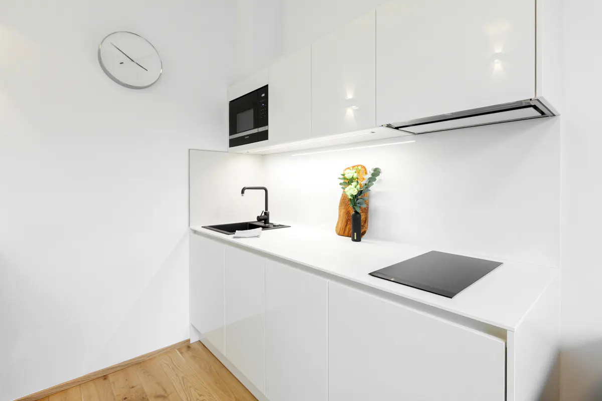 A modern, all-white kitchen with a black sink, faucet, and microwave. A clock hangs on the wall, and wood floors are visible.