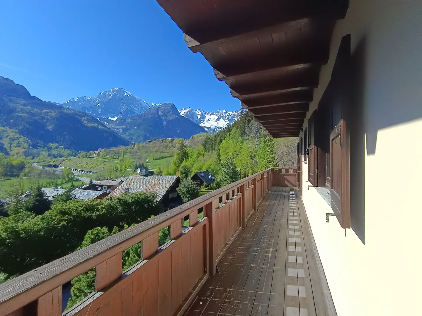 View from a wooden balcony with a brown railing, overlooking green trees, houses, and snow-capped mountains under a blue sky.