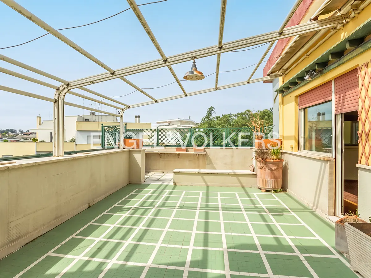 A rooftop terrace with a green and white grid floor, a metal pergola, and potted plants. A yellow building with a window is visible.