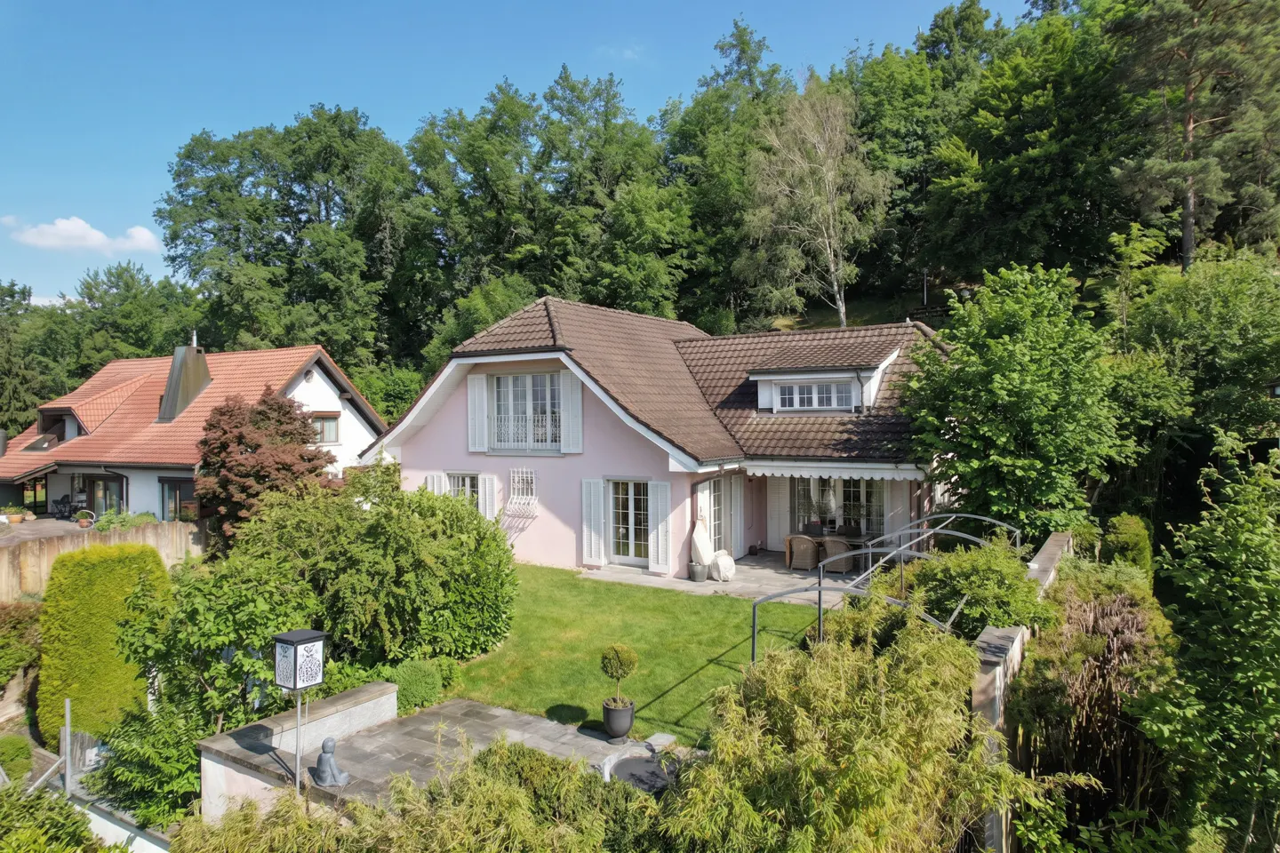 A pink two-story house with white shutters and a brown roof, surrounded by green trees and lawn.