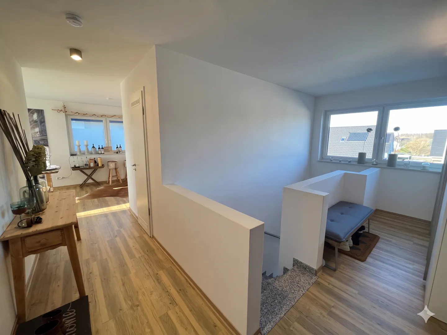 Bright hallway with wood floors, white walls, and natural light. A wooden table with a vase is on the left. Stairs lead up to a window seat.