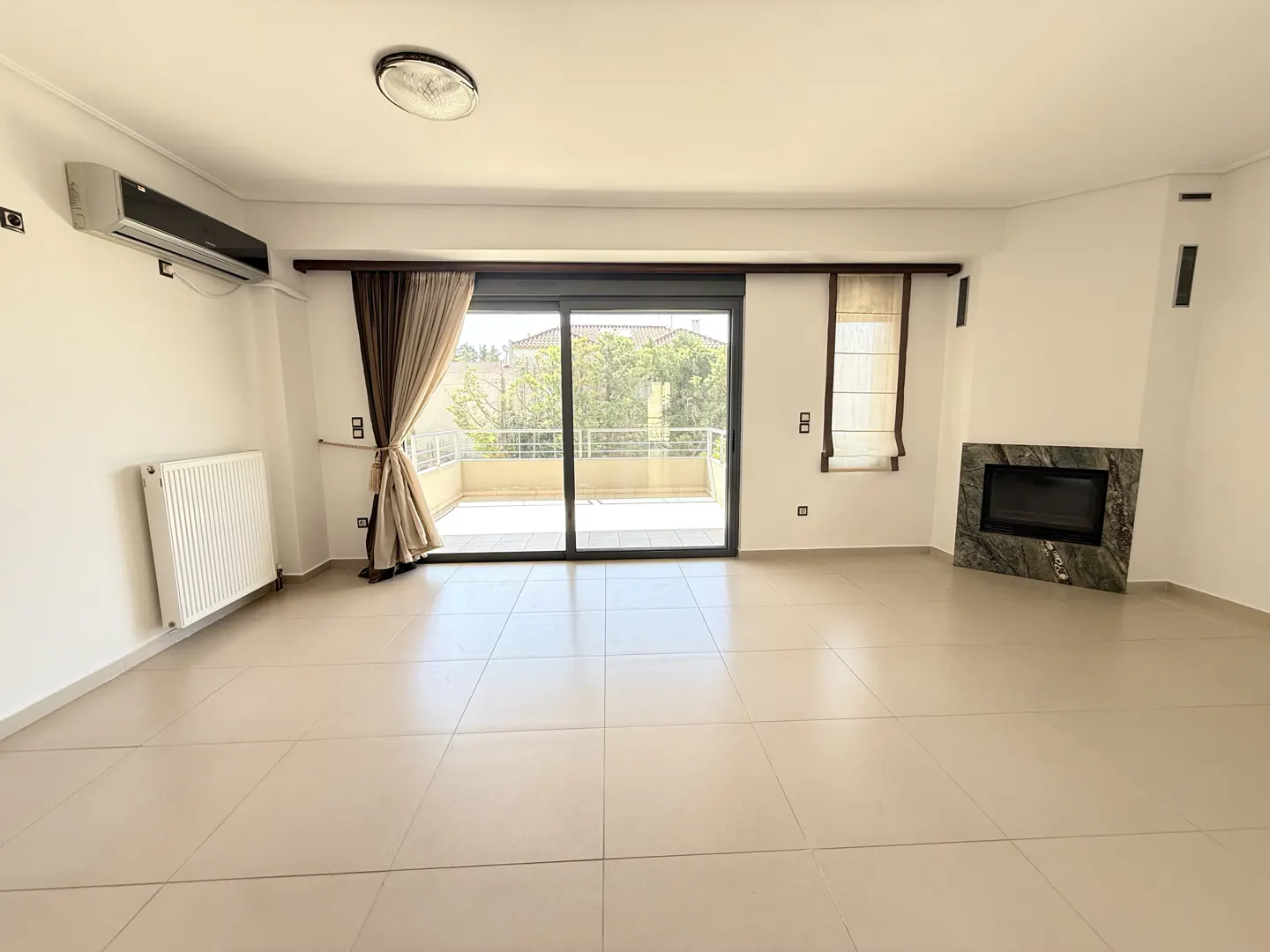 Bright, empty room with beige tile floor, white walls, and a sliding glass door to a balcony. A marble fireplace is on the right.