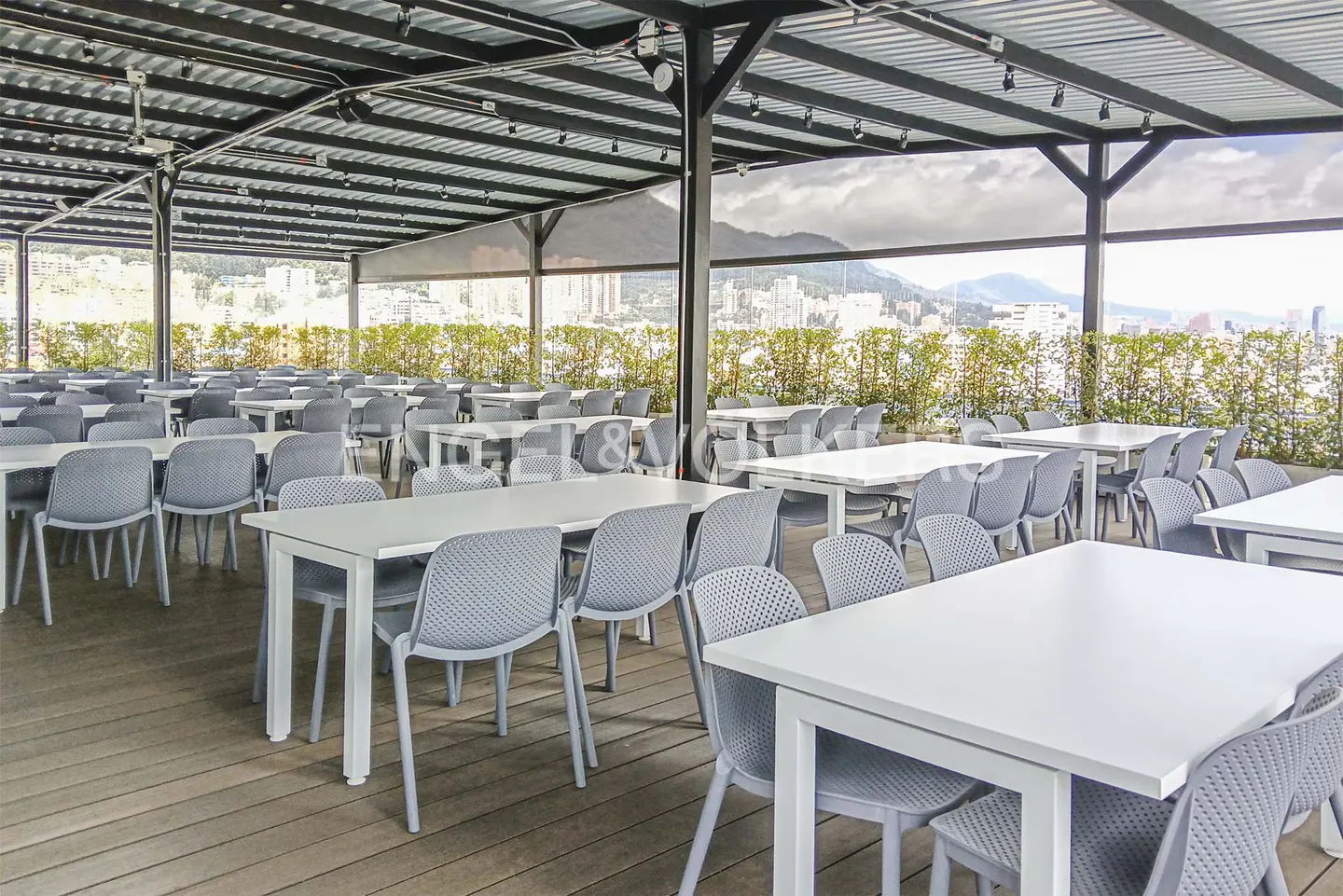 Outdoor dining area with white tables and gray chairs under a metal awning, overlooking a cityscape and green foliage.