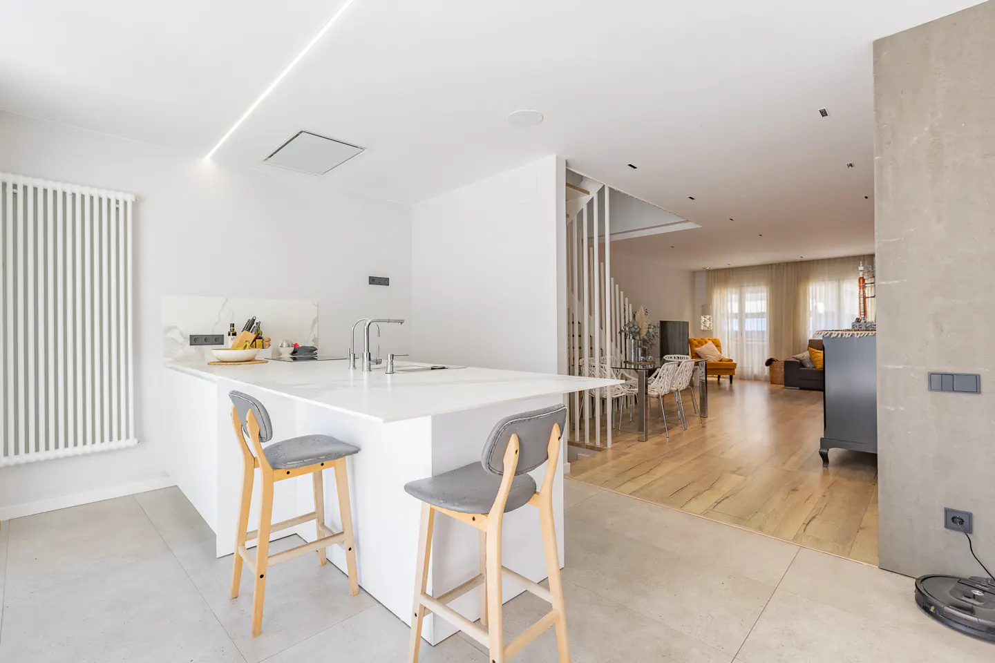 Bright, modern kitchen with white island, gray stools, and open floor plan leading to a living area with yellow armchair.