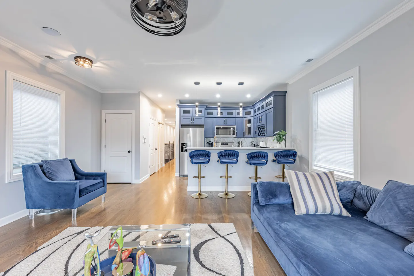 Open-concept living space with blue velvet furniture, a glass coffee table, and a kitchen with blue cabinets and gold bar stools.