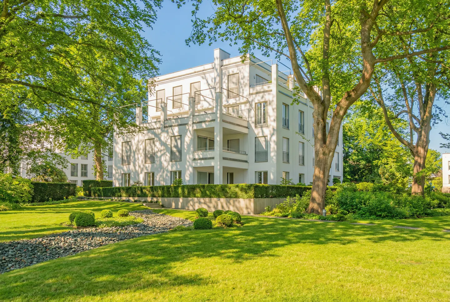 A three-story white building with balconies, surrounded by green trees and a lawn.