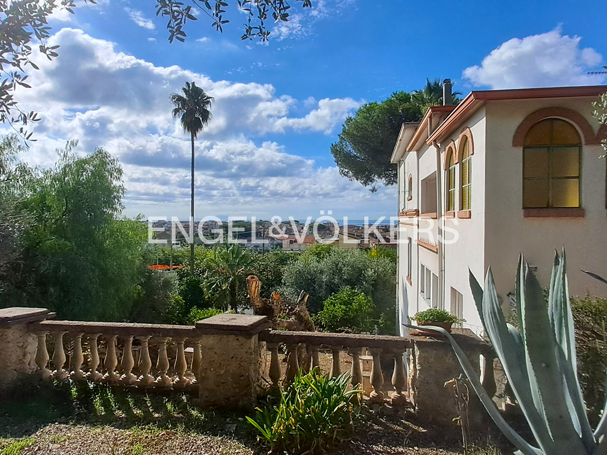 A white villa with arched windows overlooks a lush garden, palm trees, and a distant sea view under a bright blue sky.