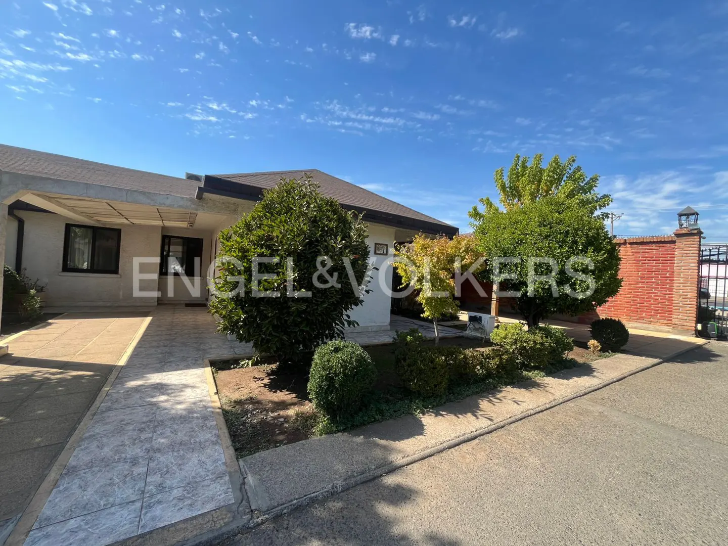 Exterior of a one-story house with a covered driveway, green trees, and a brick fence under a blue sky.