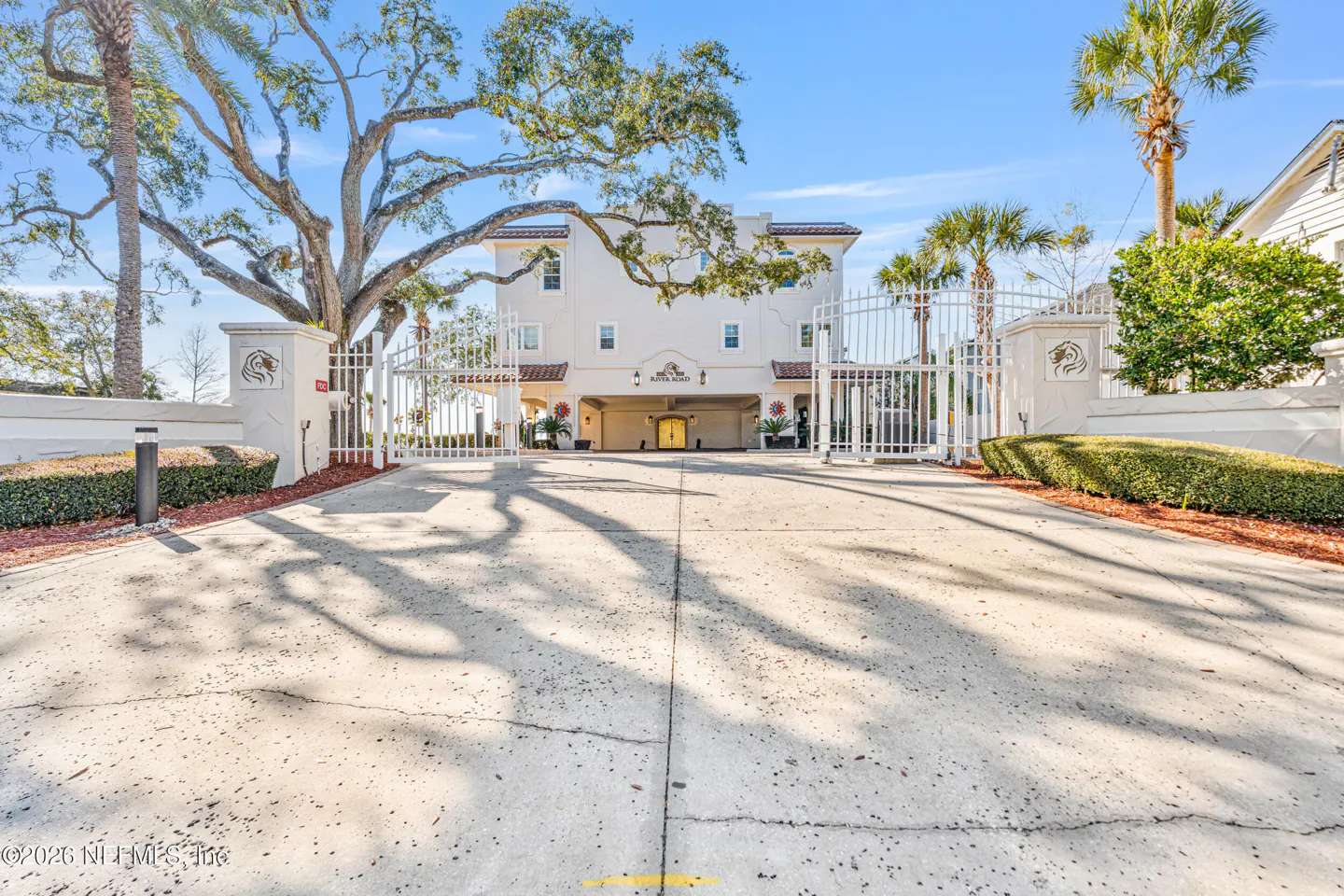 Exterior view of a gated, white, three-story building with palm trees and a large tree.
