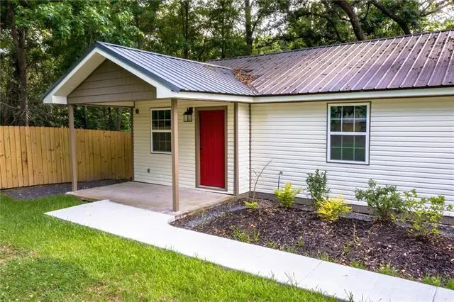 Exterior view of a one-story house with white siding, a red front door, and a metal roof. A covered porch is on the left side of the house.