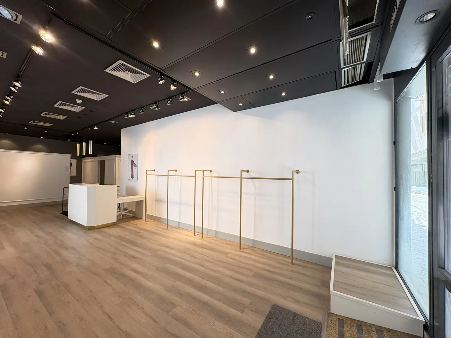Empty retail space with light wood floors, white walls, and a black ceiling with track lighting. Gold clothing racks line the wall. A white sales counter is visible.
