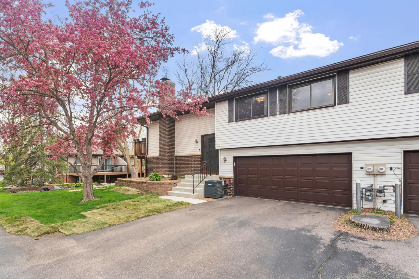 Exterior view of a two-story townhouse with white siding and a brown garage door. A pink flowering tree is on the left.