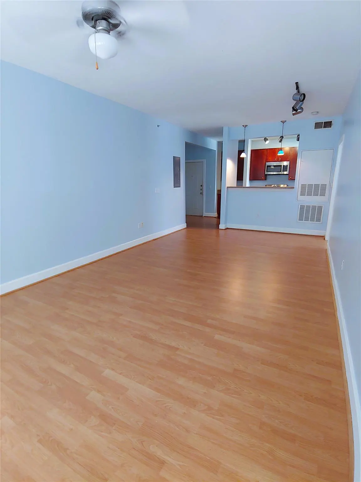 A wide shot of an empty living room with light blue walls and wood floors. A kitchen is visible in the background.