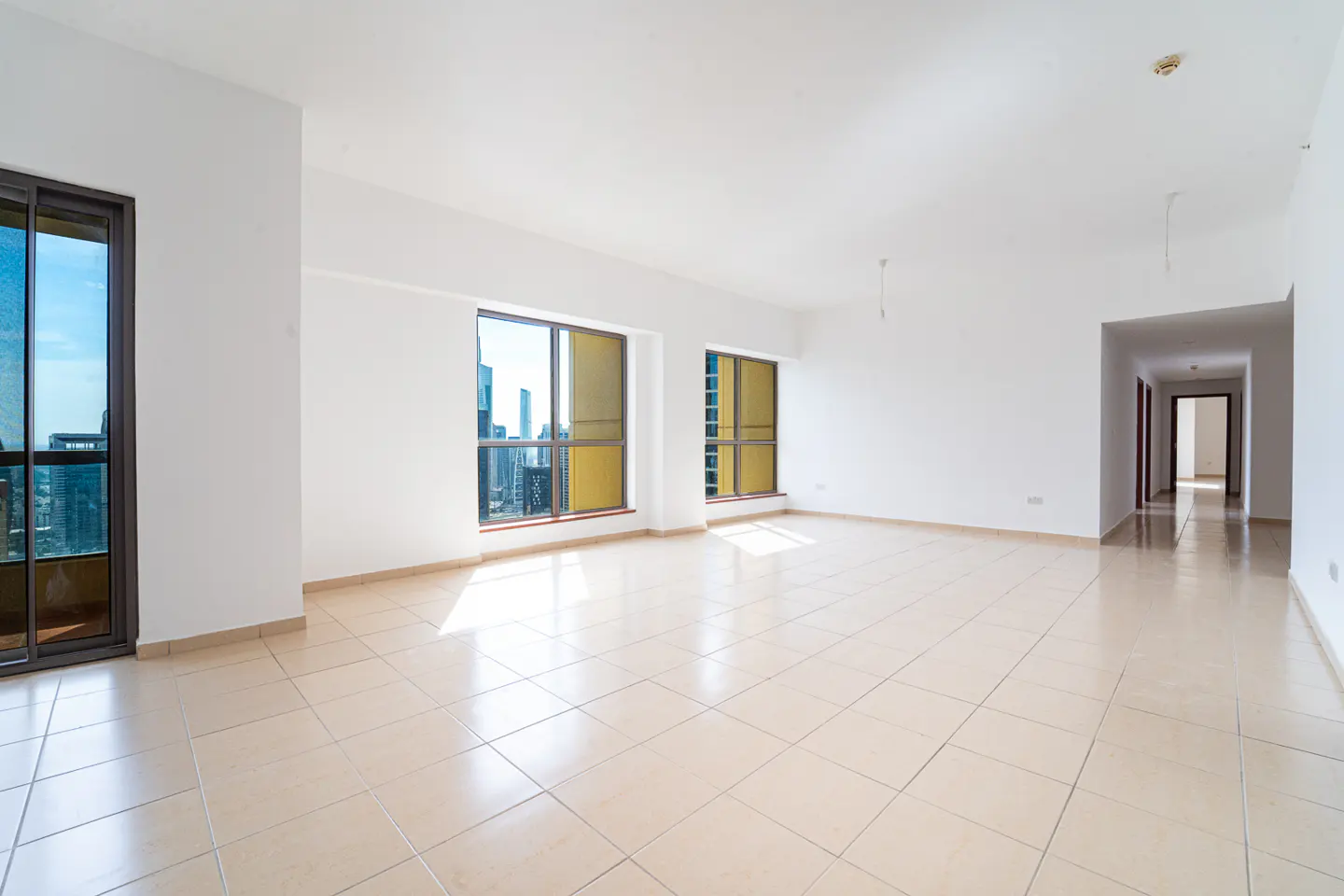 Bright, empty room with white walls, beige tile floor, and large windows showing a city view. A hallway leads to other rooms.