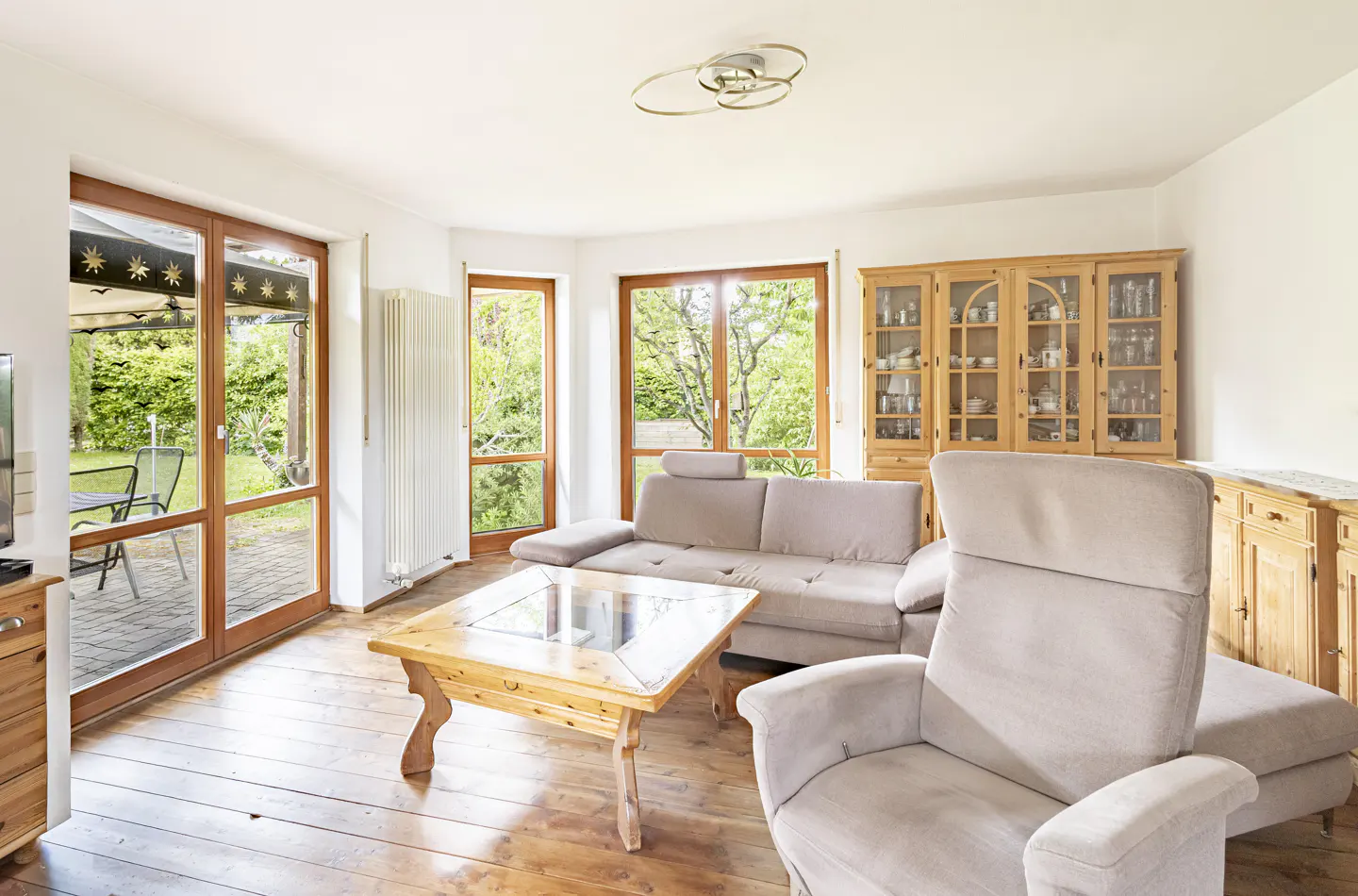 Bright living room with wood floors, beige sofa and armchair, and a wooden cabinet. Large windows overlook a green garden.