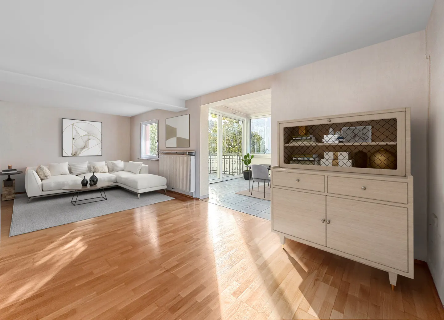 Bright living room with hardwood floors, white sectional sofa, and a light wood cabinet. A sunroom with a dining table is visible in the background.