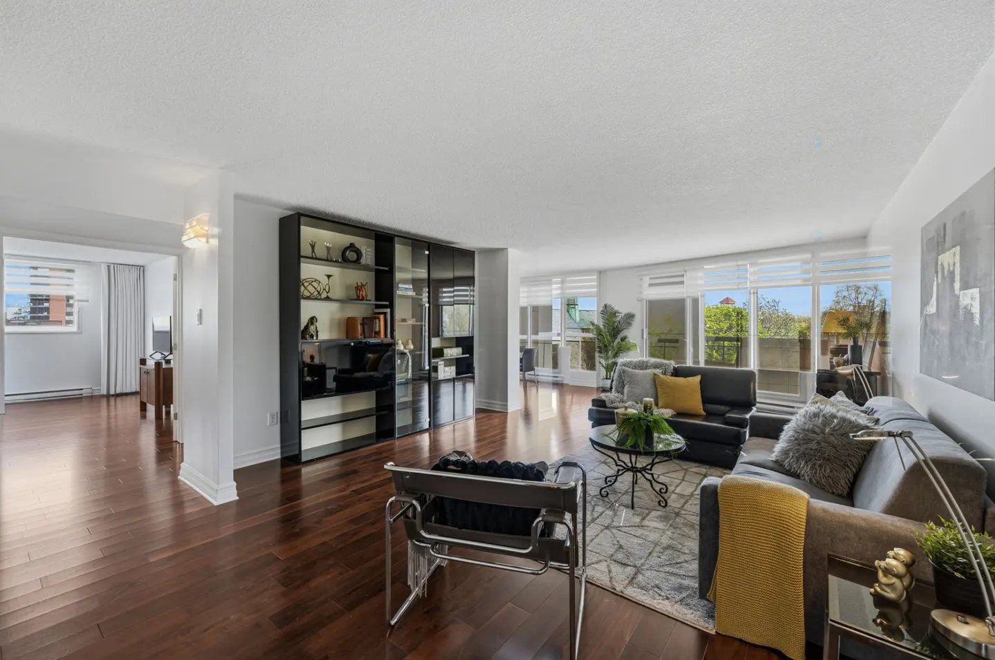 Bright living room with hardwood floors, floor-to-ceiling windows, gray sofas, and a black bookcase.
