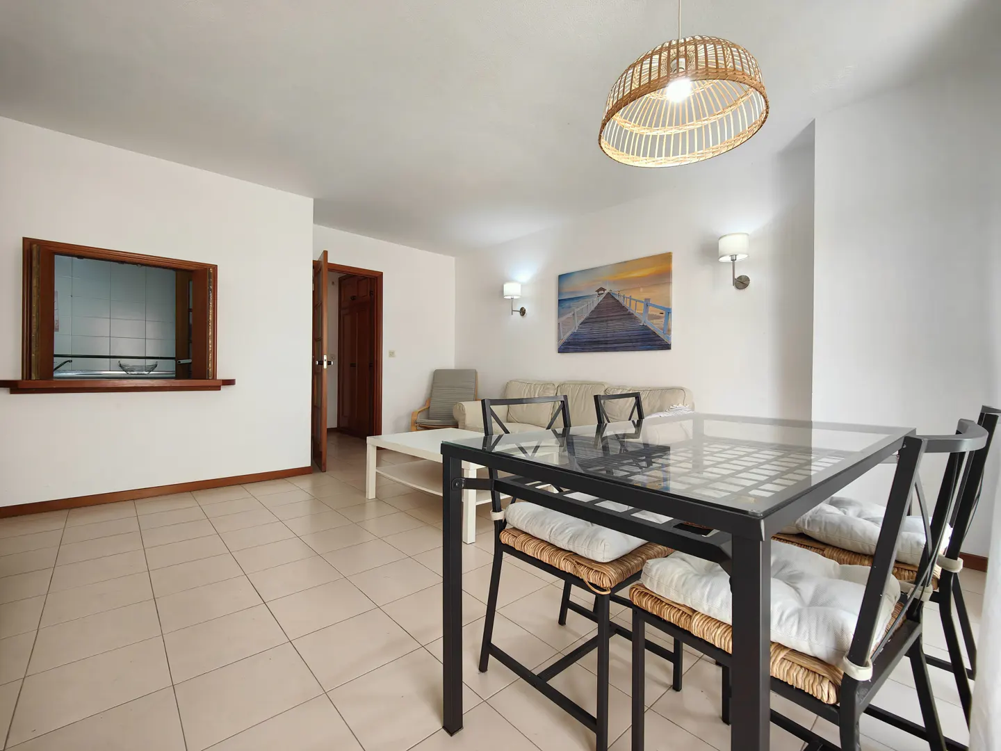 Bright living room with a glass-top table, black chairs with woven seats, and a wicker pendant light. A painting of a pier hangs on the white wall.