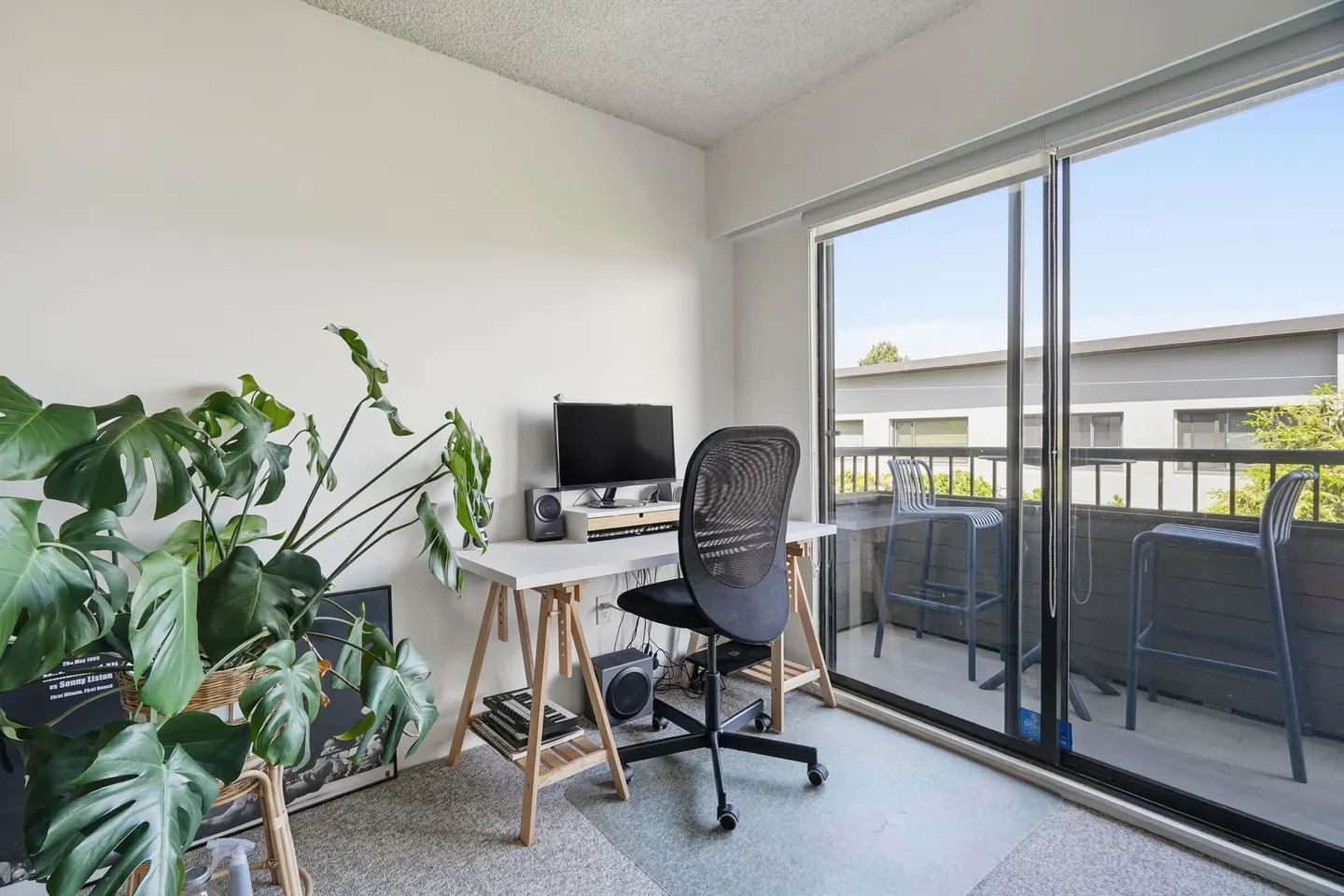 Bright home office with a white desk, black chair, computer, and a large monstera plant. Sliding glass doors lead to a balcony with bar stools.