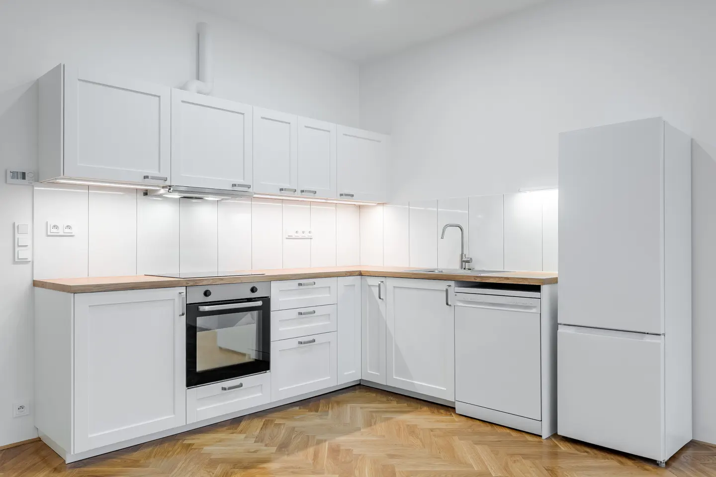 Bright, modern kitchen with white cabinets, wood countertops, and herringbone wood floor. Stainless steel appliances include oven, sink, and refrigerator.