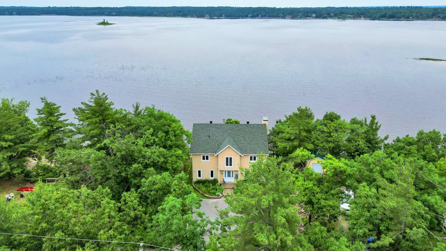 Aerial view of a beige house with a green roof, surrounded by trees, overlooking a lake.