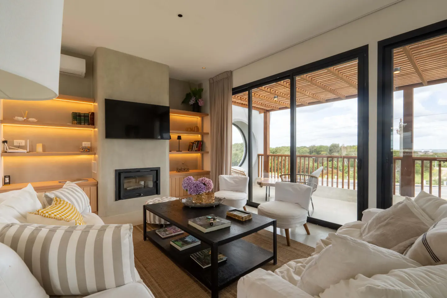 Bright living room with white sofas, a black coffee table, and a fireplace. Shelves with books and decor flank a TV. Sliding glass doors open to a balcony with outdoor seating.