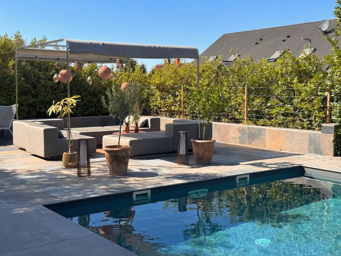 Outdoor patio with a gray sectional sofa under a canopy, potted plants, and a blue swimming pool on a sunny day.