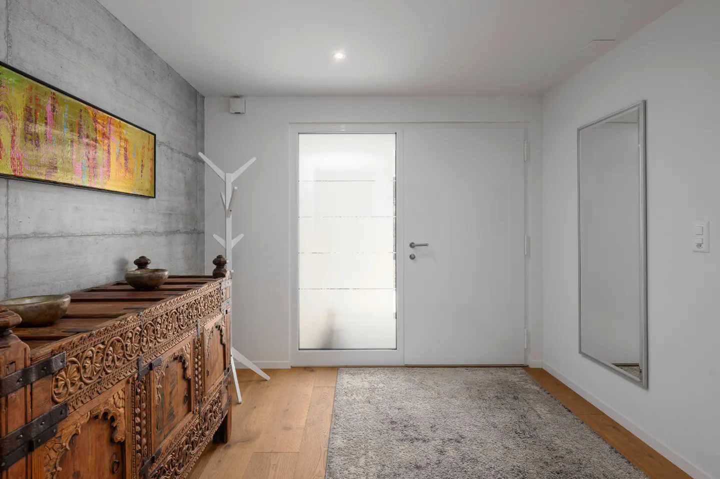 Entryway with carved wooden chest, abstract art, and white walls. A mirror and coat rack are also visible.
