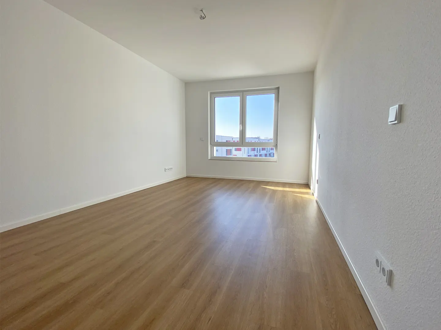 Bright, empty room with wood floor, white walls, and a window showing a blue sky and buildings.