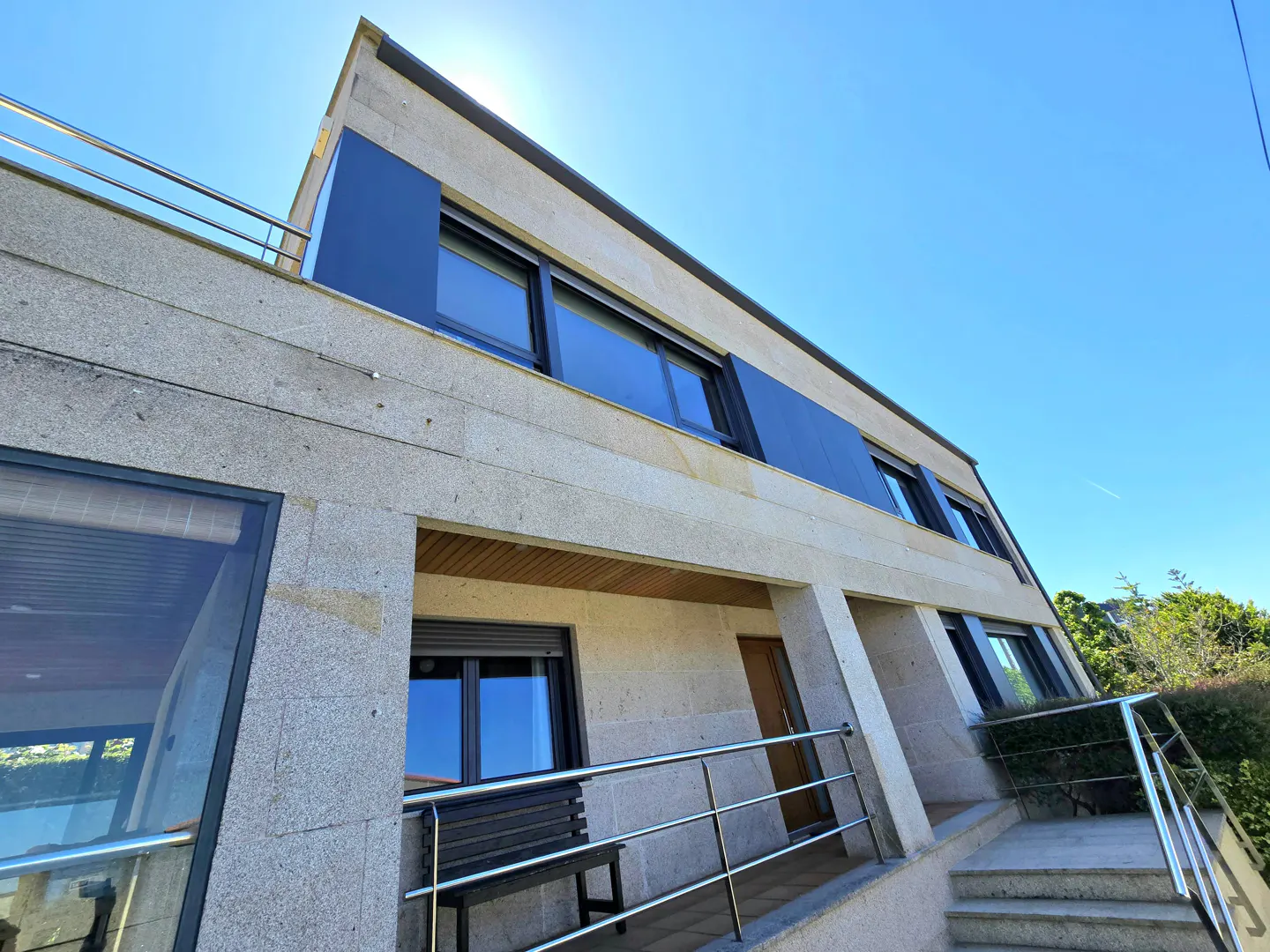 Low-angle view of a modern two-story stone house with dark window frames, a metal railing, and a wooden bench.