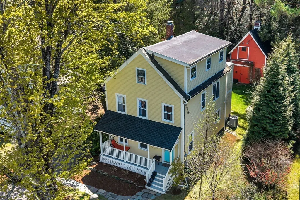 Aerial view of a yellow, two-story house with a black roof, white porch, and a blue door, surrounded by green trees.