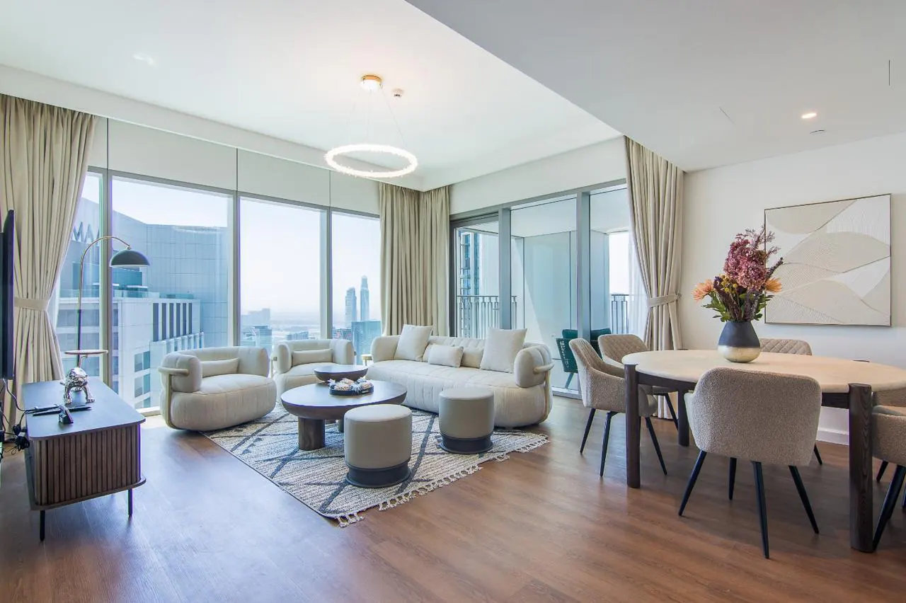 Bright, modern apartment interior with beige sofa, chairs, and dining table. Large windows offer a city view.