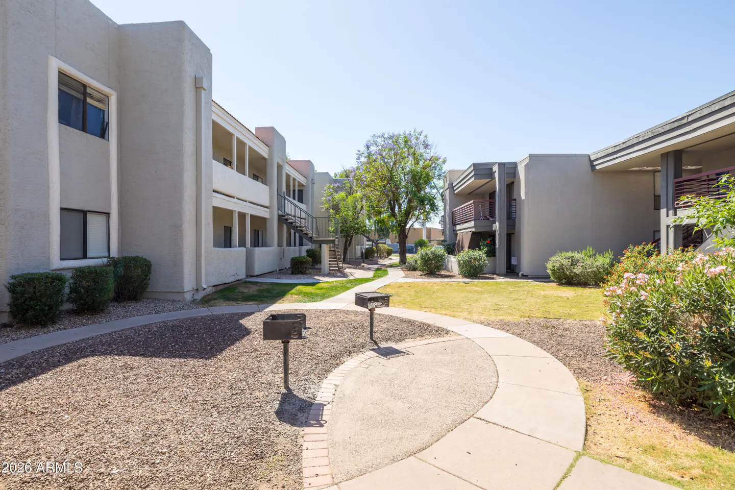 Exterior view of a light gray apartment complex with a courtyard, grills, and walkways on a sunny day.