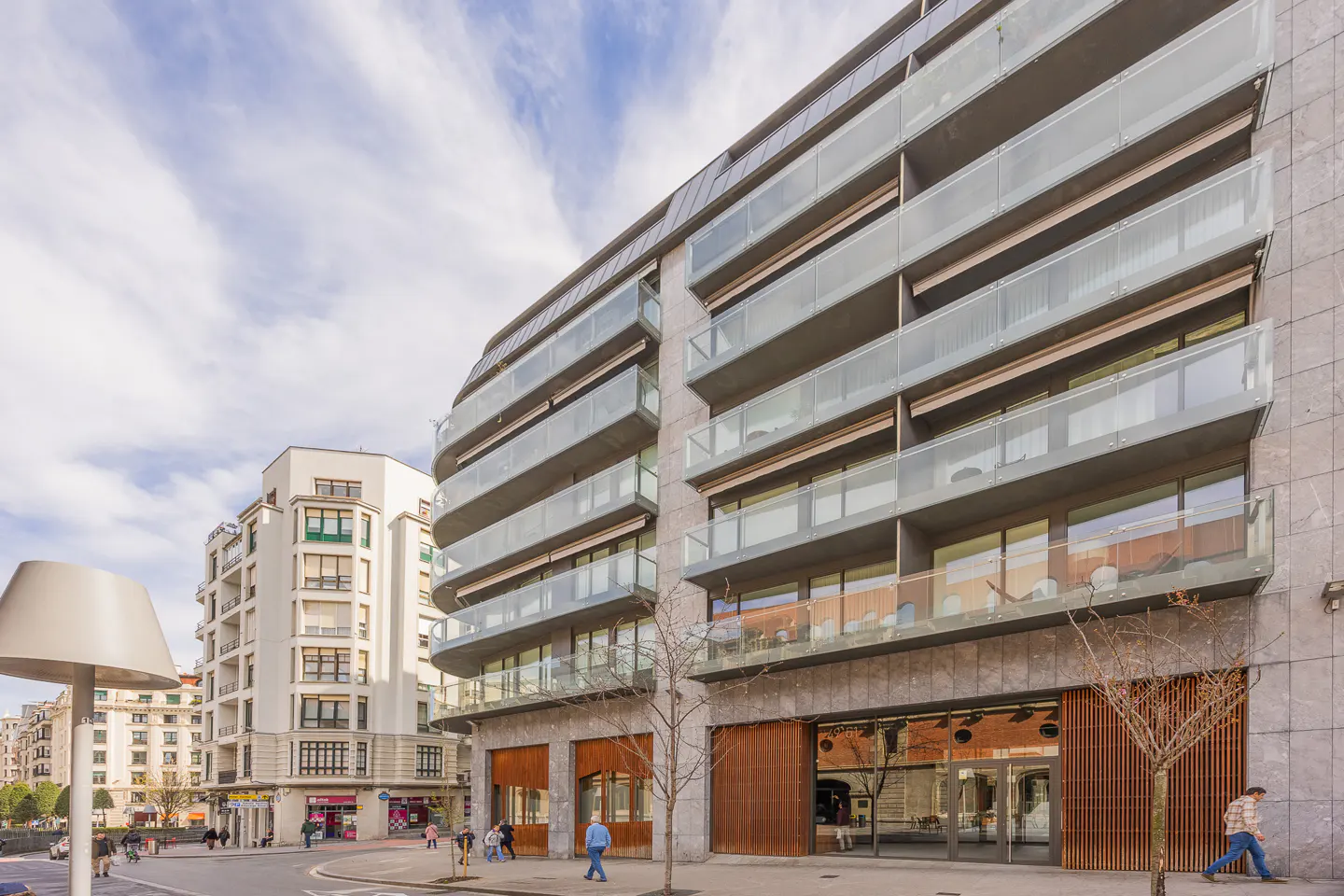 Modern apartment building with glass balconies and stone facade, street-level retail, and pedestrians on the sidewalk.