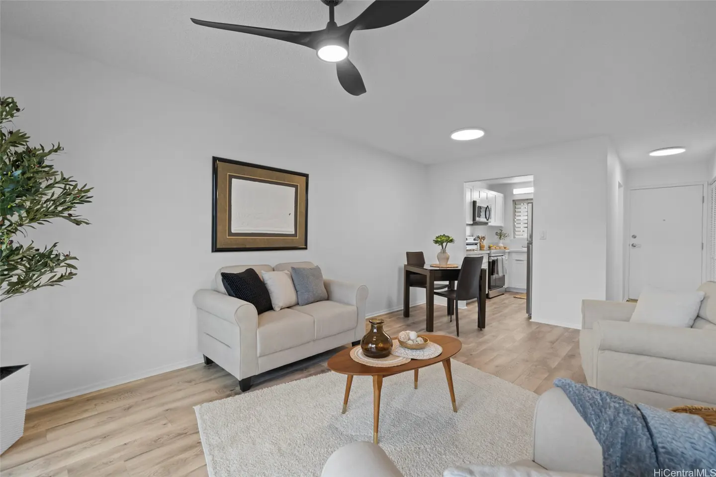 Bright living room with white walls, light wood floors, and a black ceiling fan. A cream sofa, wood table, and kitchen are visible.
