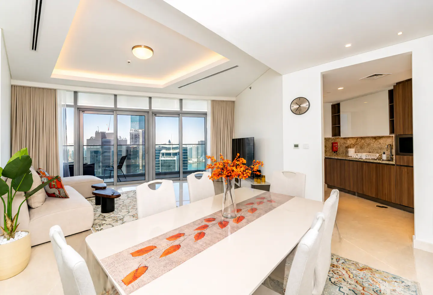 Bright, modern apartment interior with a white dining table, chairs, and a city view from the balcony. Orange flowers add a pop of color.