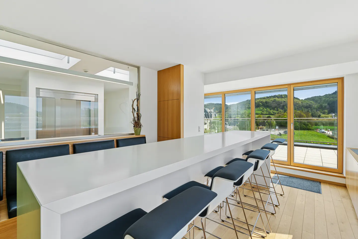 Bright, modern kitchen with a long white island and blue stools. Large windows offer a view of green hills.