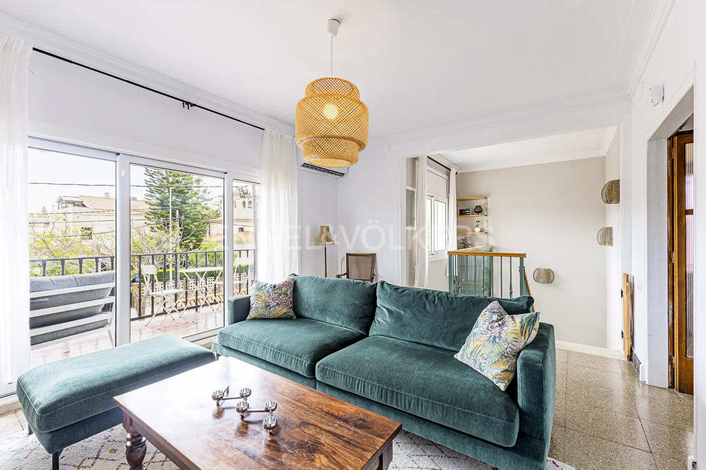 Bright living room with a green sofa, wooden table, and a balcony view. A woven pendant light hangs above.
