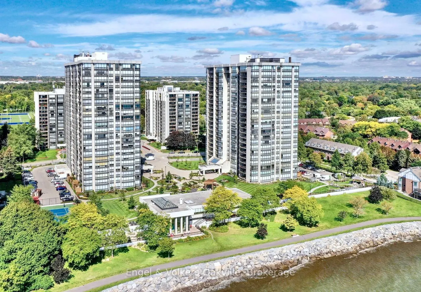 Aerial view of tall, white condo buildings with many windows, surrounded by green trees and a blue sky with white clouds. A body of water is in the foreground.