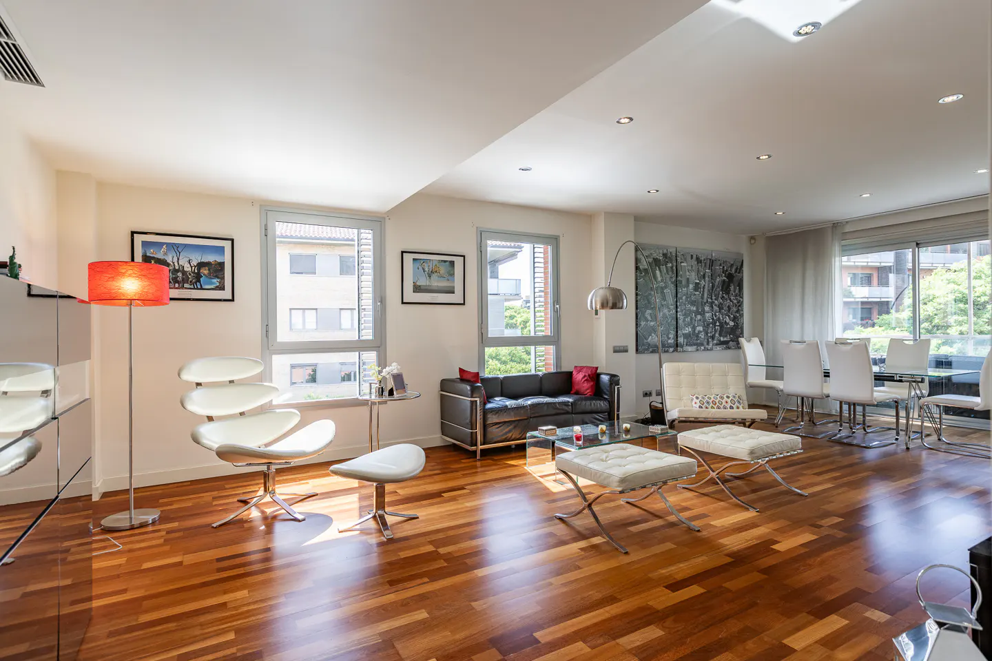 Bright, modern living room with hardwood floors, white walls, and large windows. Features a black sofa, white chairs, and a dining table.