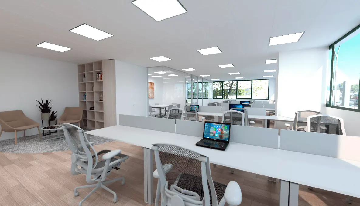 Bright, modern office space with white desks, mesh chairs, and a laptop. A lounge area with tan chairs and a bookshelf is visible.