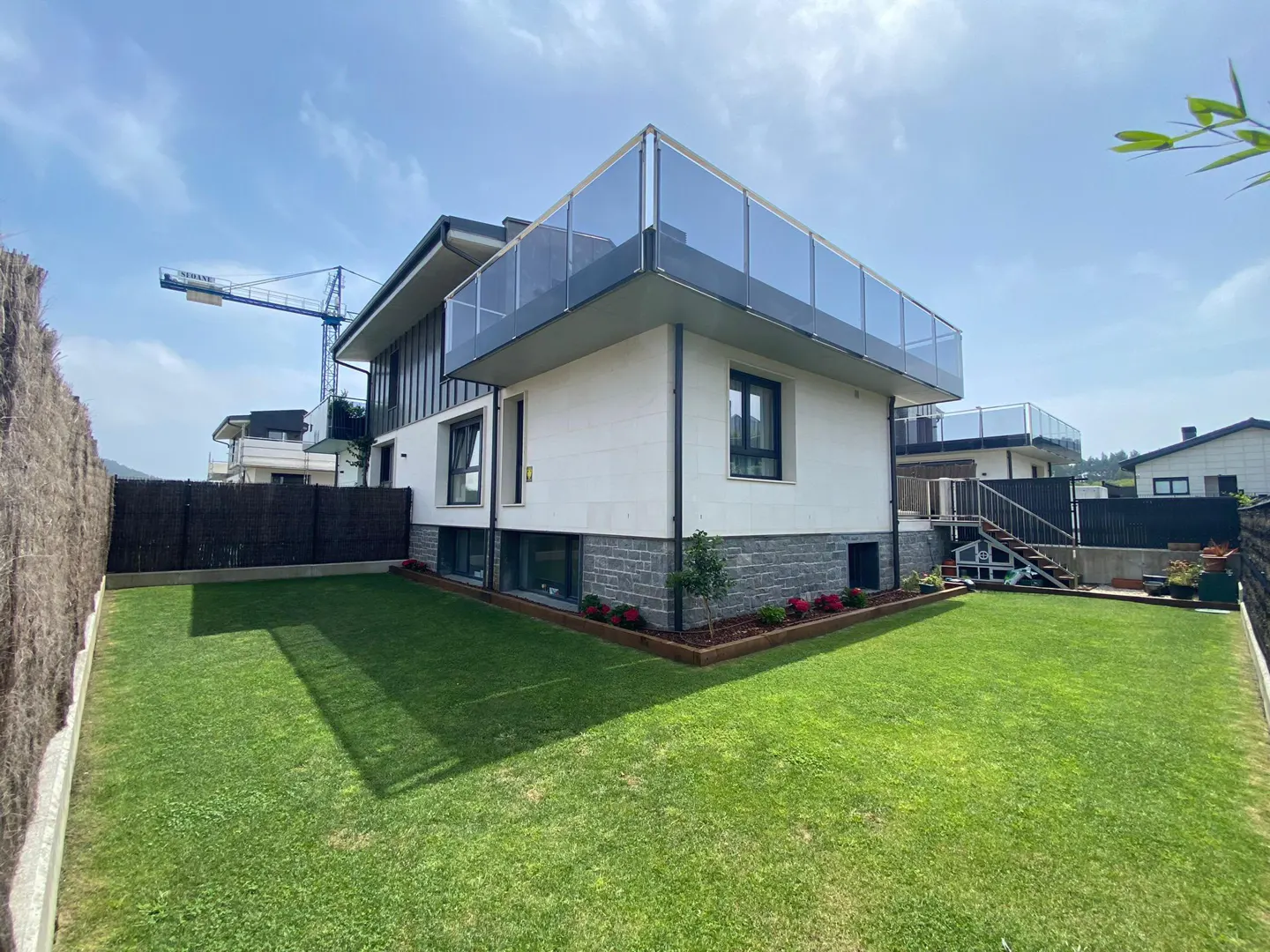Modern two-story house with a glass-railed balcony, a green lawn, and a wooden fence under a blue sky.