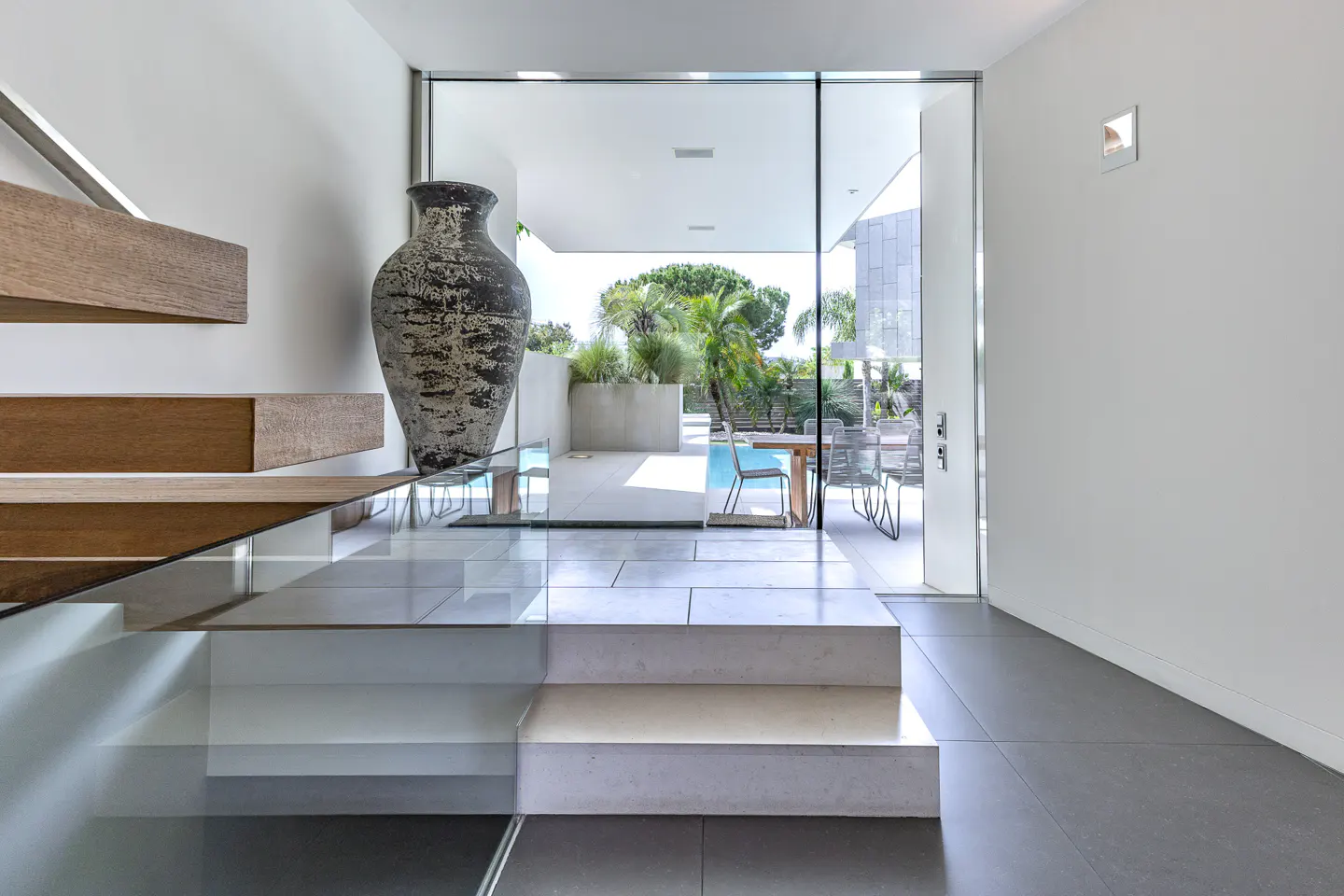 Modern home interior with floating wood stairs, glass railing, and a large decorative vase. View to a pool and patio through glass doors.